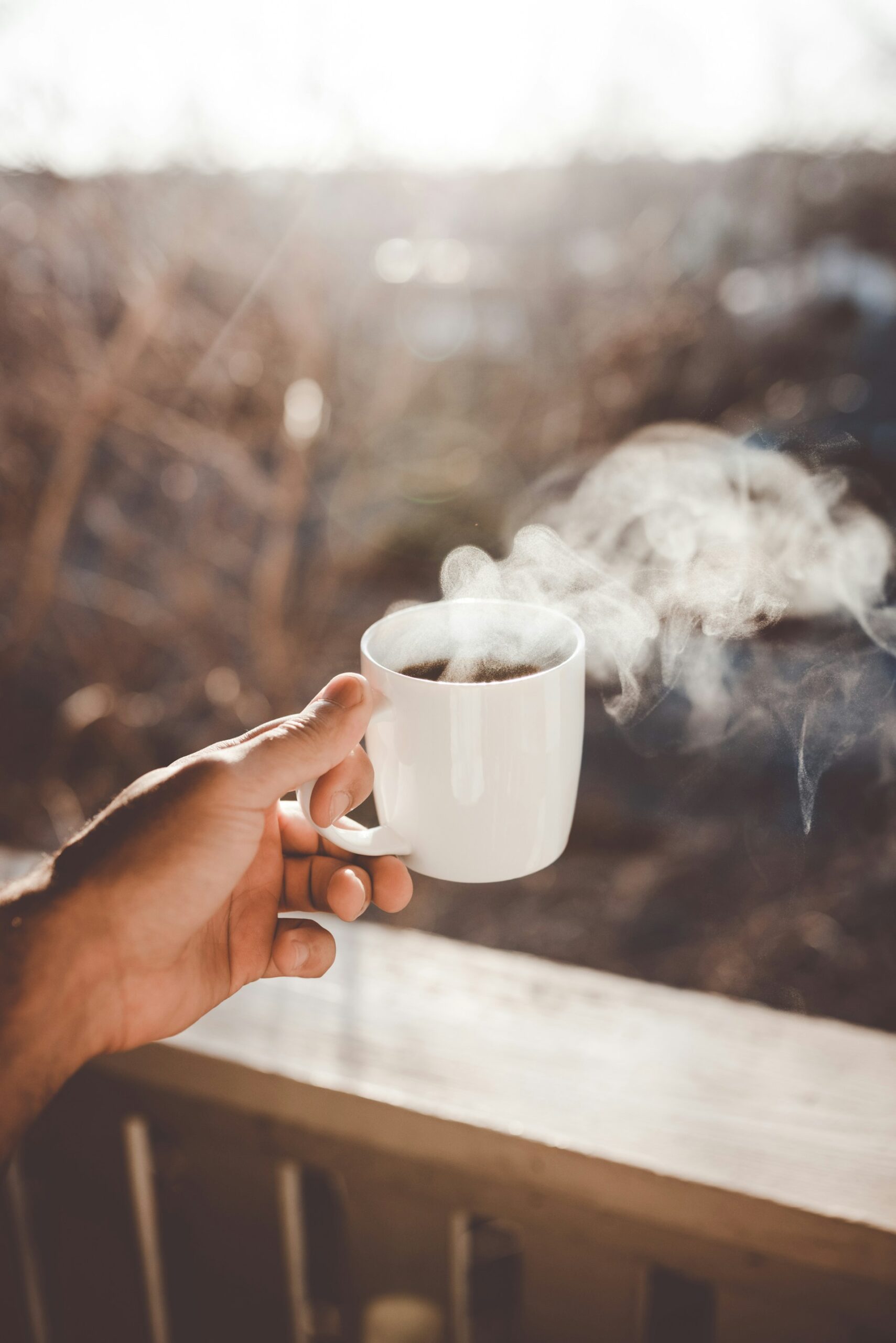 A hand holding a steaming cup of hot black coffee outdoors in soft morning light, symbolizing daily coffee consumption and heart health.