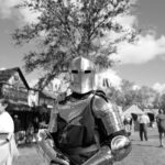 A person wearing full medieval armor stands outdoors at a historical reenactment event, symbolizing strength, protection, and readiness for battle.