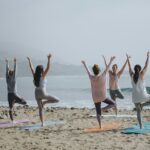 A group of women practicing yoga on the beach in tree pose, promoting heart health, stress relief, and a healthy lifestyle by the ocean.