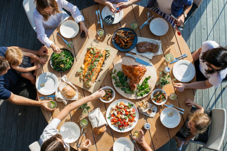 Overhead view of a family and friends sharing a relaxed outdoor meal with grilled fish, vegetables, and bread, representing balanced eating, food enjoyment, and a healthy lifestyle.