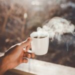 A hand holding a steaming cup of hot black coffee outdoors in soft morning light, symbolizing daily coffee consumption and heart health.