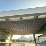Wide-angle view of the National Museum of Korea in Seoul, showing its modern architectural design with a large cantilevered roof, glass façades, and an open plaza framed by the city skyline in the background.