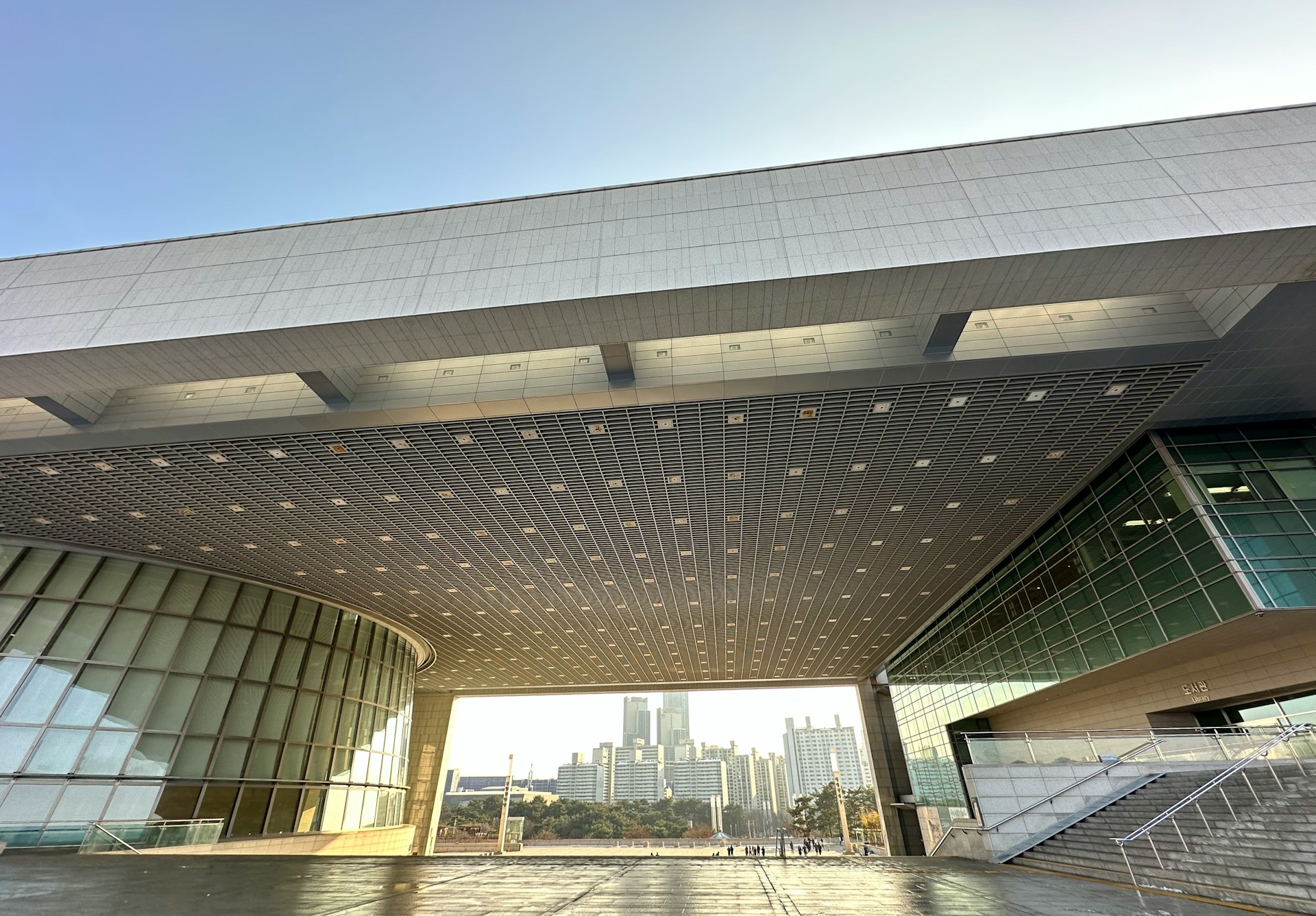Wide-angle view of the National Museum of Korea in Seoul, showing its modern architectural design with a large cantilevered roof, glass façades, and an open plaza framed by the city skyline in the background.
