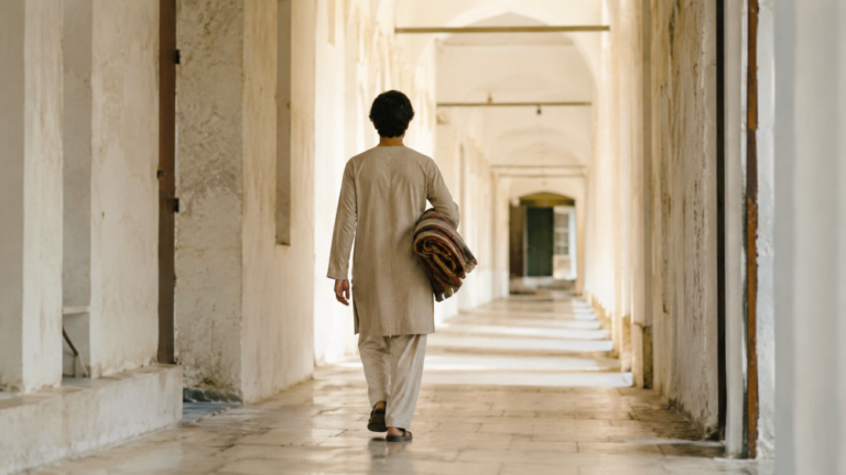 A person walks away from the camera down a long, softly lit corridor, carrying a folded blanket or mat under one arm. The scene suggests healing, purpose, and moving forward after restoration.